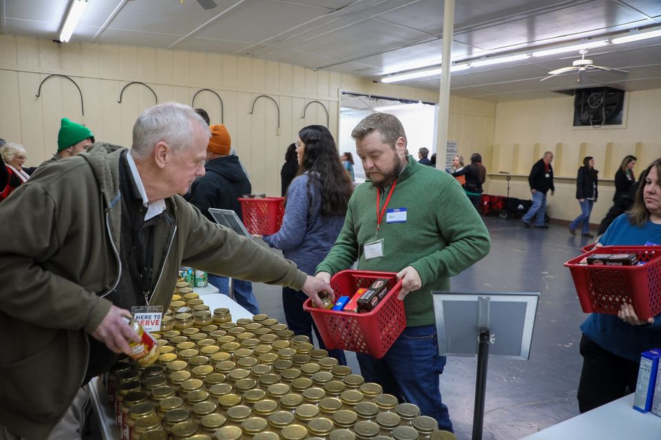 On Packing Day, a volunteer sets a jar of gravy into a red basket held by a volunteer in a green sweater, beside a table covered in jars.