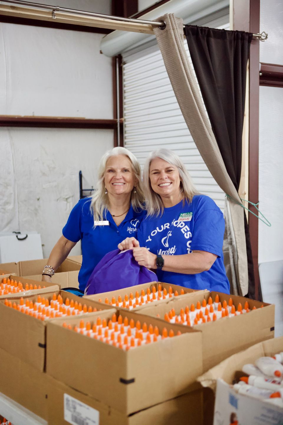 Wendy and another volunteer smile over open boxes of school glue while packing backpacks.