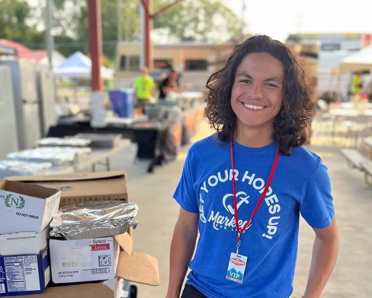 A smiling volunteer in a Your Hopes Up shirt beside boxes of meals
