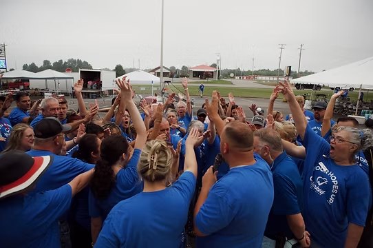 Volunteers in blue Marked For Life shirts gathered in a circle with hands raised in prayer at an outdoor event