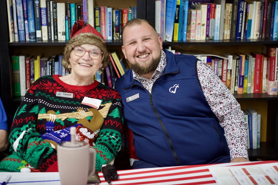 Carmen, in a Christmas sweater and Santa hat, and Logan LeGrande smile at a Christmas Blessings check-in table.