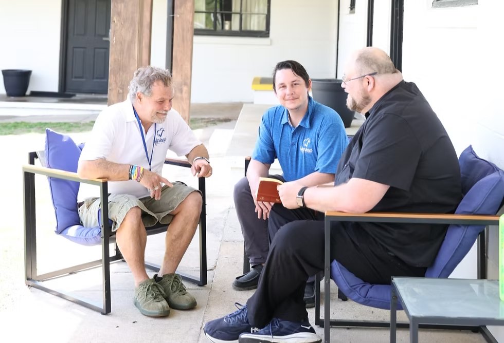 Three men talking together on a porch, one holding a Bible